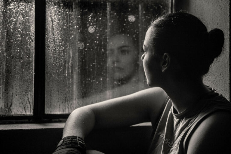 Nachdenklicher Blick aus dem Fenster bei Regen. Spiegelbild einer jungen Frau im Fensterglas. Gitter vor dem Fenster. Schwarzweiß